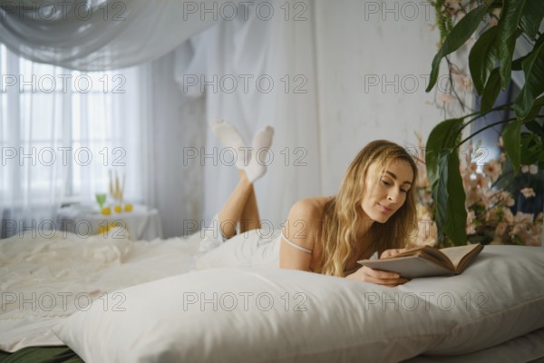 A woman is lying on a bed, reading a book with a relaxed pose. The room is bright and has plants