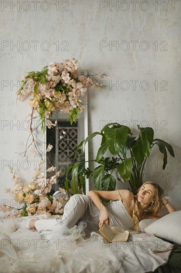 A woman relaxes on a bed while reading a book in a sunny room. She is surrounded by plants and