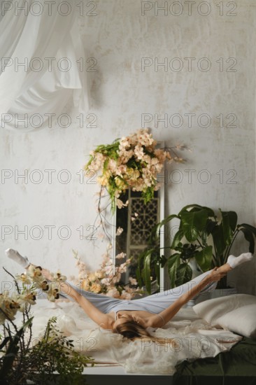 Woman in pajamas stretches on a bed in a bright room filled with plants and flowers. The stretching