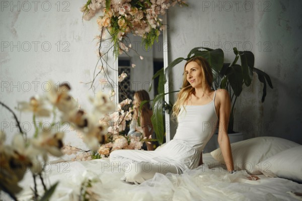 A woman sits on a bed in a bedroom decorated with flowers and plants. Natural light fills the