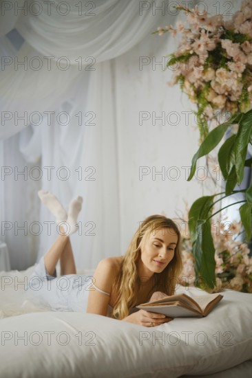 Reading a book while lying on a bed in a room with flowers and a plant during afternoon light