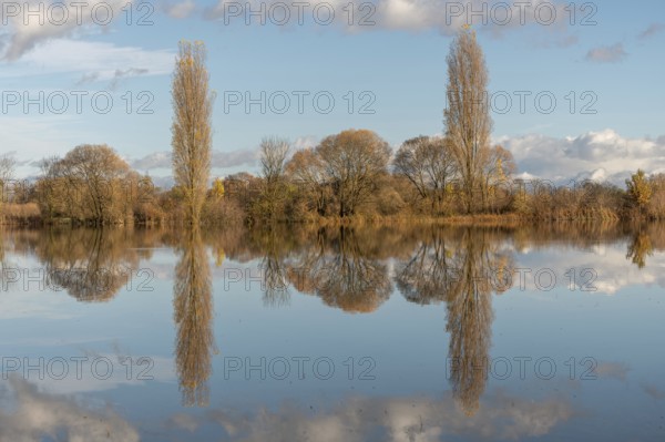The river flows flat and reflects clouds and mountains under a clear sky. Autumn trees draw the