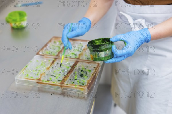 A person in a blue glove paints with a brush from a green paint pot on shapes, vegan chocolate