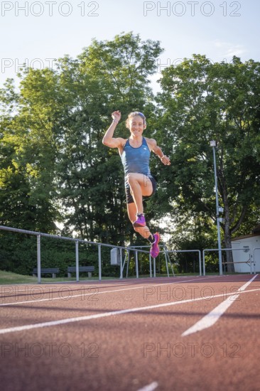 A person wearing blue sportswear jumps with raised arm on the running track in sunshine, middle
