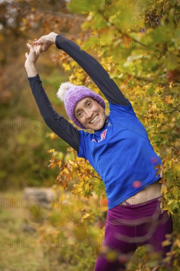 Woman doing stretching exercises in autumn forest, smiling happily surrounded by colorful leaves,
