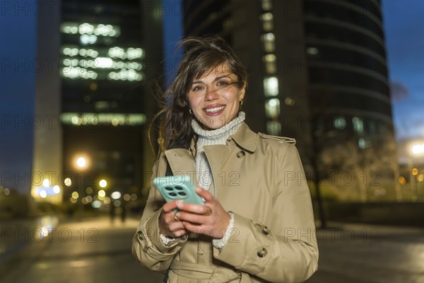 Young woman in a trench coat stands on a lit city street at dusk, smiling at the camera while