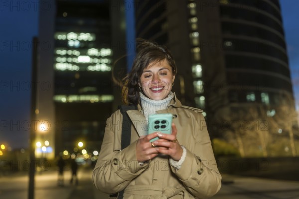 Young woman smiling while walking through the city at night, engaging with her mobile phone and