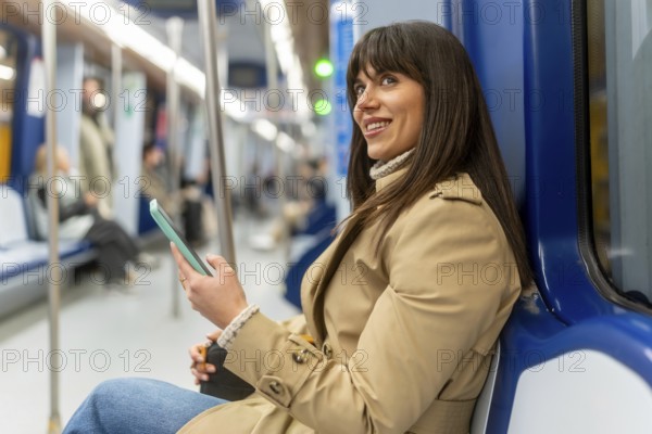 Young woman sits in a subway car smiling at her phone while commuting through the city, relaxed and