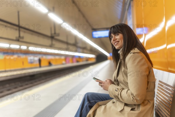 Young woman commuting, checking her mobile phone and smiling while sitting on a bench at an empty
