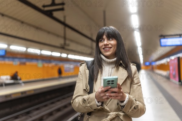 Young woman standing on a subway station platform, looking away and smiling while checking her