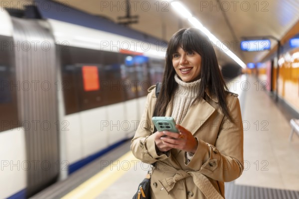 Young woman standing on a subway platform, wearing a trench coat and sweater, happily interacting
