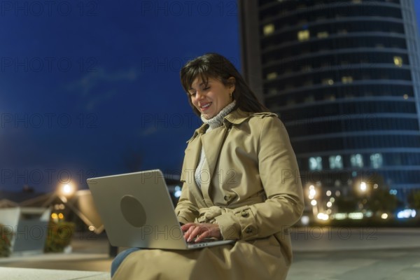 Woman in a trench coat smiling and typing on a laptop, connecting to the internet and managing her