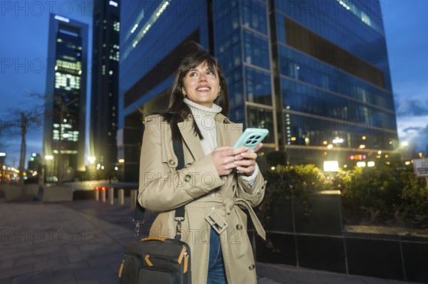 Young professional woman walking along a city sidewalk at dusk, holding a smartphone and carrying a