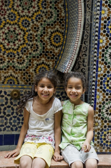 Two smiling girls in front of a wall with colorful tiles, alley in the old town, Souk, Fez El Bali,