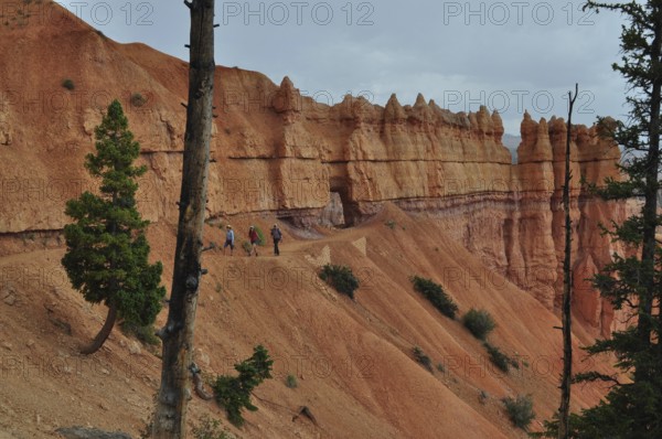 Hikers on a trail along dramatic red cliffs under looming clouds, Bryce Canyon National Park, Utah,