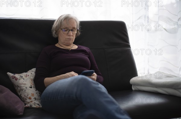 Elderly woman with glasses, gray hair, sitting on sofa and using smartphone, cell phone, Stuttgart,