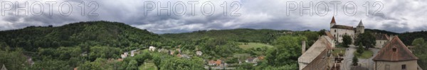 Panoramic view of a hilly landscape with a castle complex and thick greenery under cloudy sky,