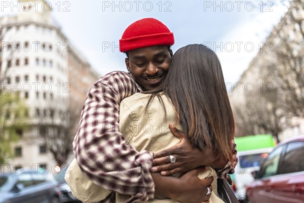 Young man in a red beanie smiles as he embraces his partner on a city street, sharing a warm,