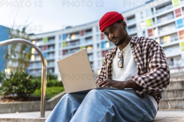 Young black man sitting on urban steps, focused on his laptop, working or studying remotely in an