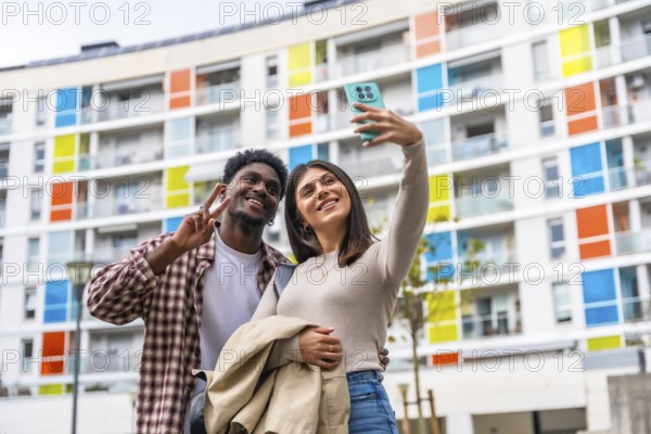 Happy diverse couple smiling and posing for a smartphone selfie, showing a peace sign while