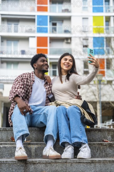 Happy diverse young couple sitting on concrete steps, smiling and taking a cheerful selfie together