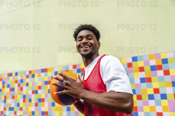 Young man smiling and holding a vibrant basketball in sportswear, standing against a colorful tiled