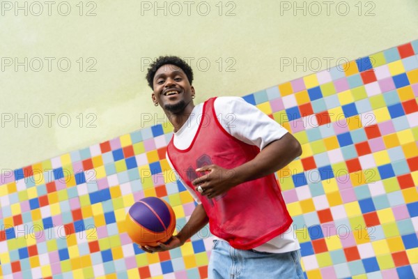 Young black man happily holding a basketball, dressed in a red sports vest and jeans, posing