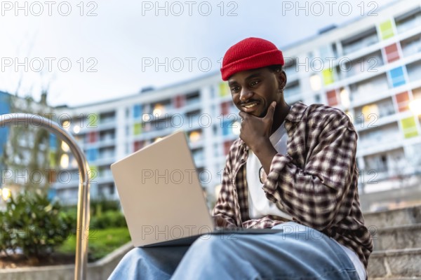 Young black man sitting on apartment steps with laptop, smiling and thoughtful as he works or