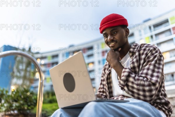 Young black man smiling while working on a laptop computer with casual clothes and a red beanie,