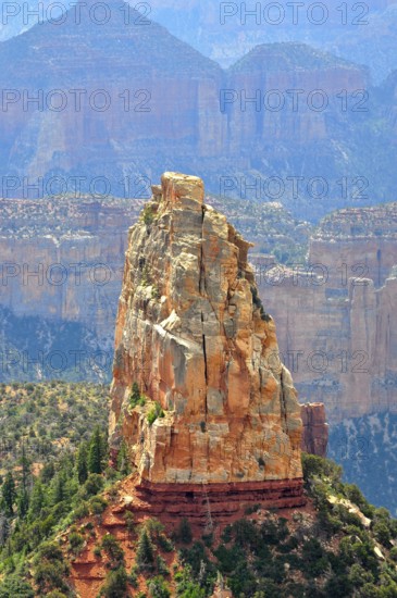 A massive rock rises steeply in a canyon landscape, surrounded by other rocks and sparse