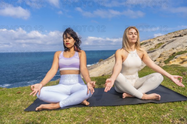 Two healthy multiracial women performing yoga and meditation poses in lotus position on a yoga mat