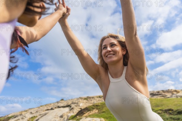 Two smiling women stretching their arms to the sky, connecting their hands in a yoga pose outdoors,