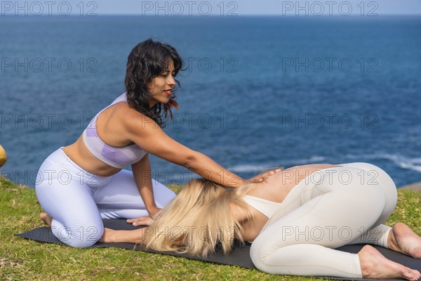 Instructor guiding woman in child's pose on grassy seaside during outdoor yoga session, promoting