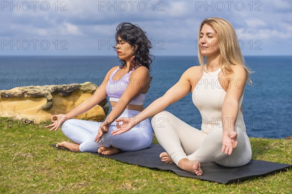 Two diverse women sitting in lotus pose on yoga mats by the ocean, finding tranquility and balance
