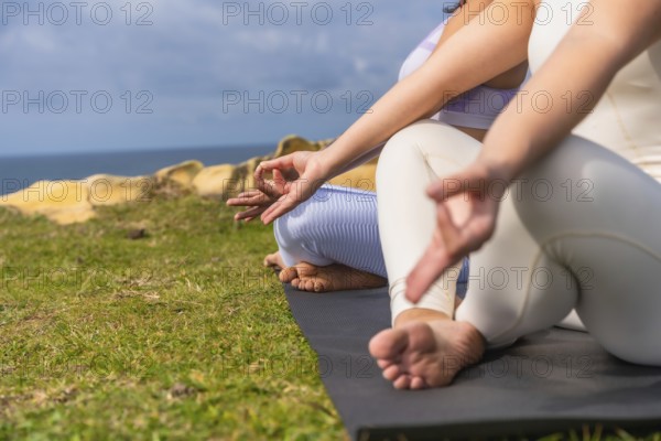 Two women practicing yoga and meditating outdoors on yoga mats, sitting in lotus pose with hands in