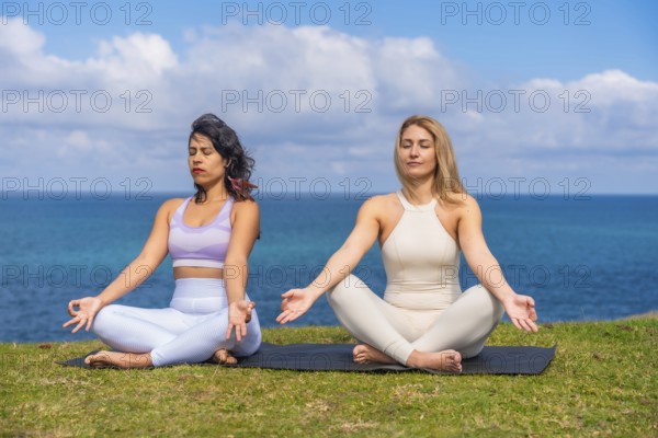 Two women sit cross legged on a mat by the ocean, practicing yoga and meditation together at