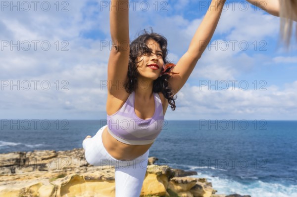 Woman balancing in a yoga warrior pose, stretching arms towards the sky on a cliff overlooking the
