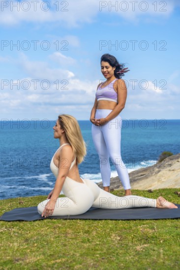 Two women stretching on a yoga mat by the ocean while practicing yoga and meditation, enjoying a