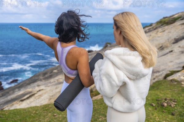 Two women standing together on a grassy cliff overlooking the ocean, one raising an arm and holding