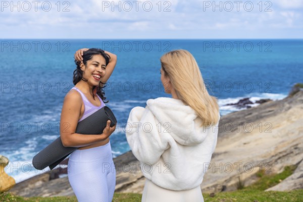 Two women friends standing on a cliff overlooking the ocean, one holding a yoga mat and laughing