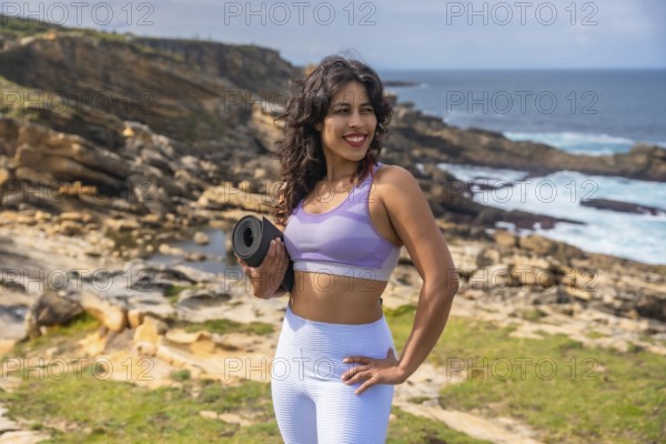 Young woman standing on coastal cliffs holding a yoga mat, smiling, looking away, preparing for an