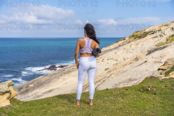 Woman standing on a grassy cliff edge, holding a yoga mat and looking out at the vast blue ocean,