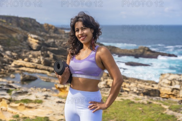 Woman standing on a rocky coastline holding a yoga mat, preparing for an outdoor fitness session