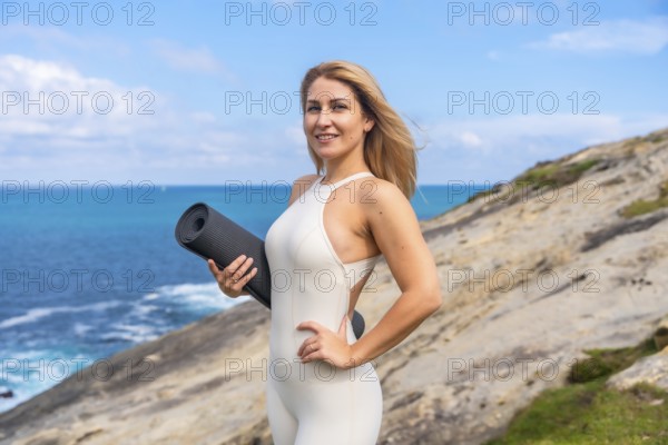 Woman standing confidently on a coastal cliff with a yoga mat, embracing a healthy and balanced