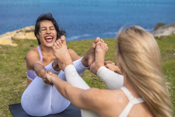 Two women performing a yoga pose together on a mat on green grass by the sea, connecting through
