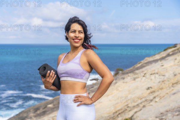 Woman smiling, holding a rolled yoga mat on a rocky cliff, looking towards the bright blue ocean,