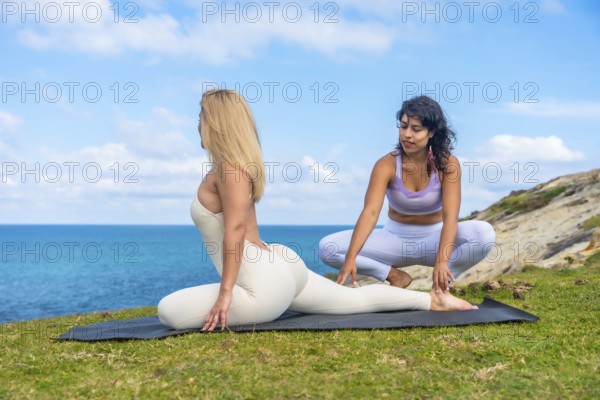 Two women are practicing yoga on a mat outdoors, with one instructor supporting a student in a