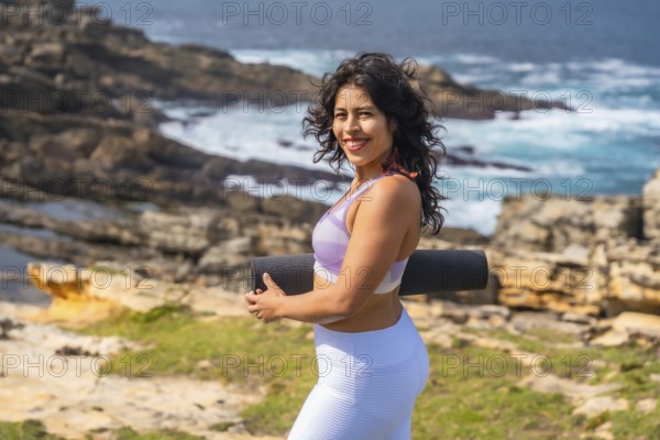 Woman smiling and carrying a rolled yoga mat or exercise mat, standing on a rocky coastline with