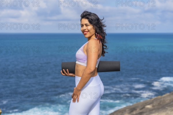 Woman smiling and holding a black yoga mat, standing on a cliff edge with the vast blue ocean and