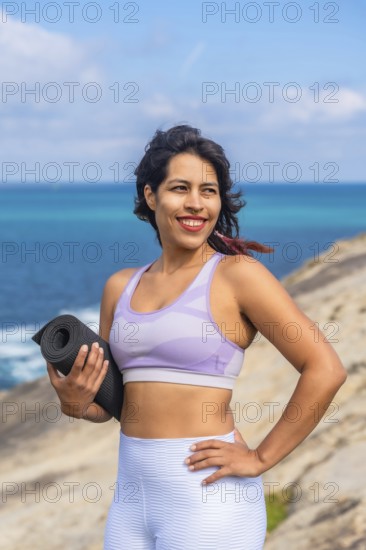 Woman holding a yoga mat and smiling, standing on a rocky cliff overlooking the blue ocean during